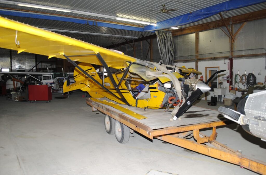 The recovered yellow Avid Magnum airplane wreckage on a trailer inside a hangar. The fuselage is crushed, the nose and engine compartment are severely damaged, and the left wing is buckled. The propeller shows damage consistent with impact forces.